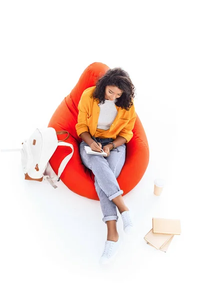Visão de alto ângulo da adolescente afro-americana estudante estudando enquanto sentado em saco de feijão isolado no branco — Fotografia de Stock