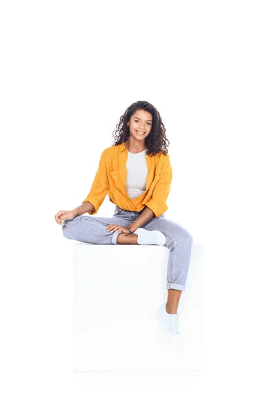 Teenage african american student girl sitting on white cube and looking at camera isolated on white — Stock Photo