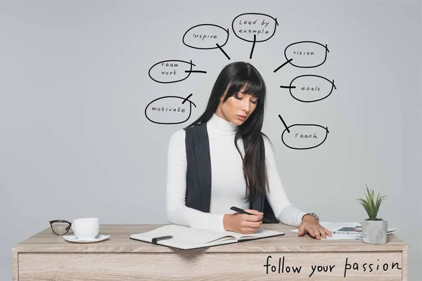 Attractive brunette businesswoman writing something at table isolated on gray with speech bubbles and follow your passion inscription — Stock Photo