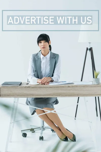 Красива бізнес-леді, сидячи за столом і дивлячись на камеру на сірому з рекламою з нами напис — стокове фото