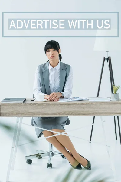 Beautiful businesswoman sitting at table and looking at camera on gray with advertise with us inscription — Stock Photo