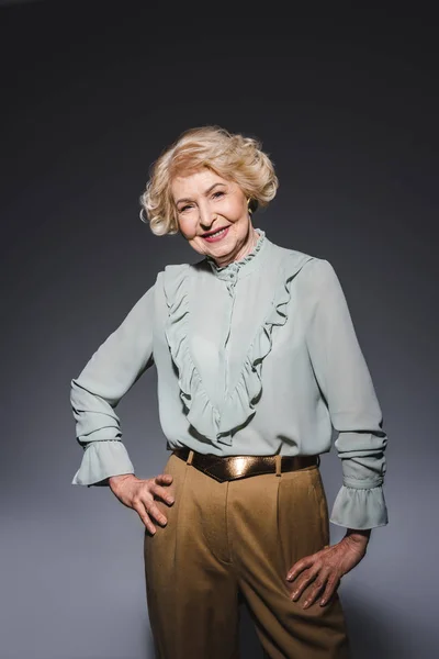 Mujer mayor sonriente en camisa elegante mirando a la cámara en gris oscuro - foto de stock