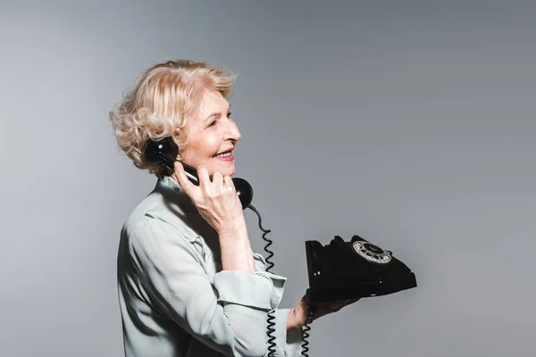 Vista laterale della donna anziana sorridente che parla dal telefono vintage isolato sul grigio — Foto stock