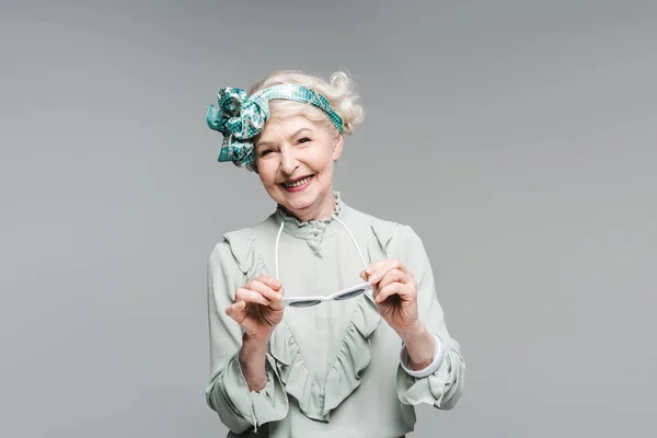 Щаслива старша жінка в стильному вінтажному одязі та сонцезахисних окулярах ізольована на сірому — стокове фото