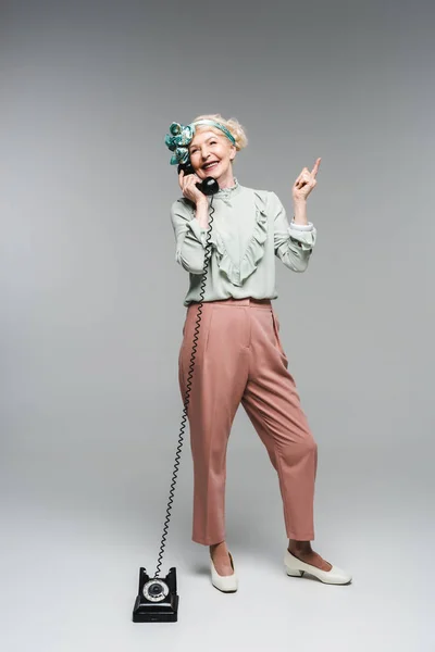 Smiling senior woman talking by vintage phone and pointing up on grey — Stock Photo