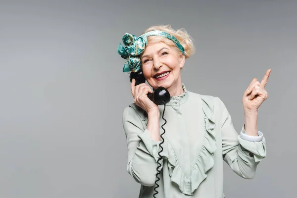 Happy senior woman talking by vintage phone and pointing up isolated on grey — Stock Photo