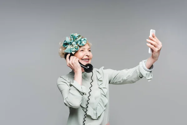 Усміхнена старша жінка бере селфі, розмовляючи вінтажним телефоном ізольовано на сірому — стокове фото