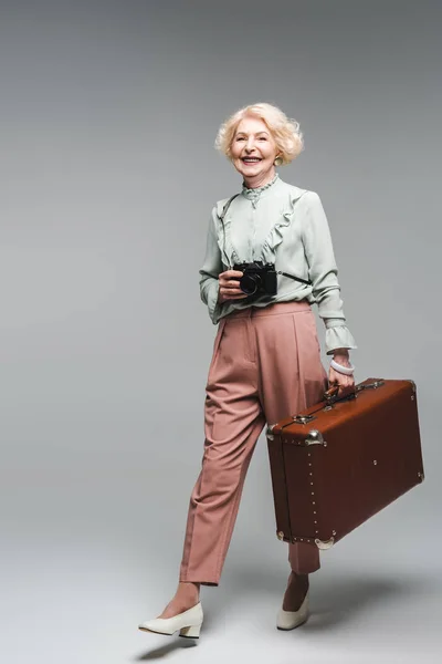 Elegante mujer mayor caminando con la maleta vintage y cámara de cine en gris - foto de stock