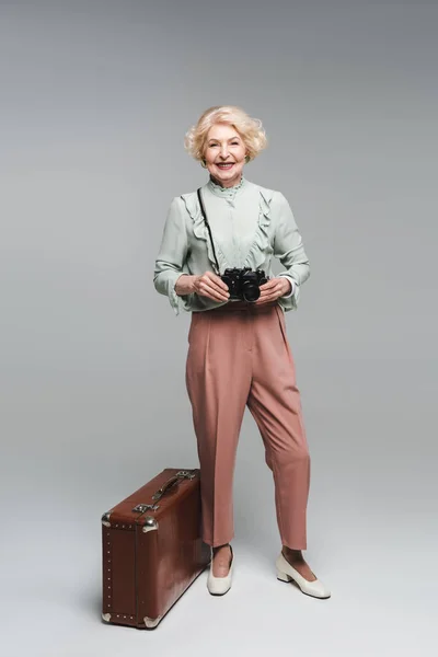 Beautiful senior woman with vintage suitcase and film camera on grey — Stock Photo