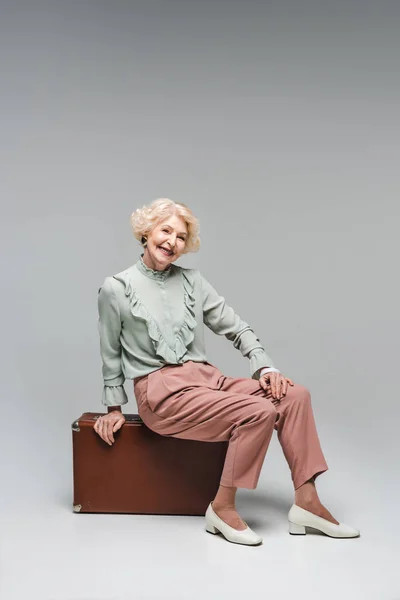 Belle femme âgée assise sur une valise vintage sur gris — Stock Photo