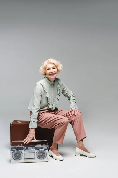 Beautiful senior woman with boombox sitting on vintage suitcase on grey — Stock Photo