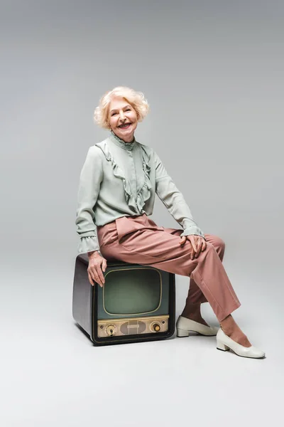 Schöne ältere Frau sitzt auf Vintage-TV auf grau — Stockfoto