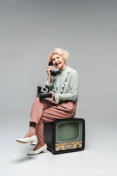 Beautiful senior woman talking by rotary phone while sitting on vintage tv on grey — Stock Photo