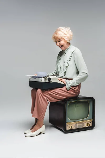Schöne Seniorin mit Schreibmaschine, während sie im Vintage-Fernseher auf grau sitzt — Stockfoto