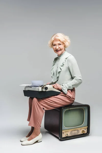 Mujer mayor sonriente usando máquina de escribir mientras está sentado en la televisión vintage en gris — Stock Photo