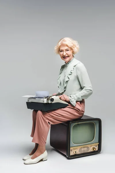 Smiling senior woman using typewriter while sitting on vintage tv on grey — Stock Photo