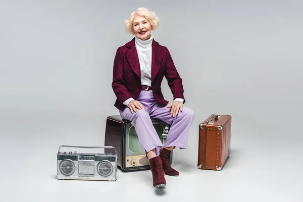 Attractive senior woman sitting on vintage tv on grey — Stock Photo