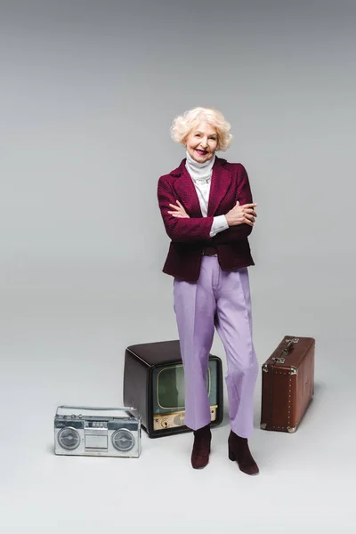 Beautiful senior woman with crossed arms standing in front of vintage tv, boombox and suitcase on grey — Stock Photo