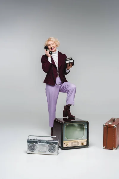 Красива старша жінка розмовляє по телефону, стоячи на старовинному телевізорі на сірому — стокове фото