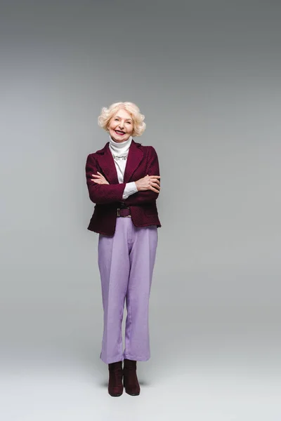 Donna anziana sorridente con le braccia incrociate in abiti alla moda guardando la fotocamera sul grigio — Foto stock