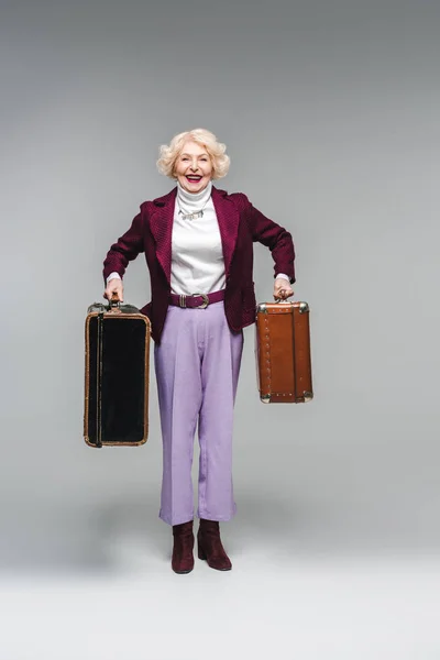 Happy senior woman holding vintage suitcases and looking at camera on grey — Stock Photo
