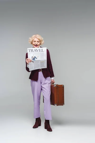 Beautiful senior woman holding suitcase and reading travel newspaper on grey — Stock Photo