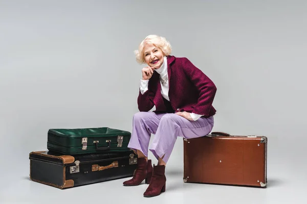 Schöne Seniorin sitzt auf Vintage-Koffer auf grau — Stockfoto