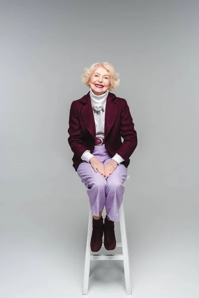Красива старша жінка в стильному одязі, сидячи на стільці на сірому — стокове фото
