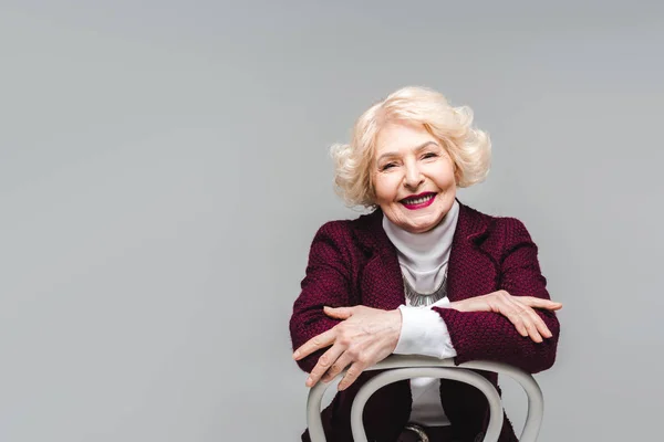 Mujer mayor sonriente sentada en la silla y mirando a la cámara aislada en gris - foto de stock