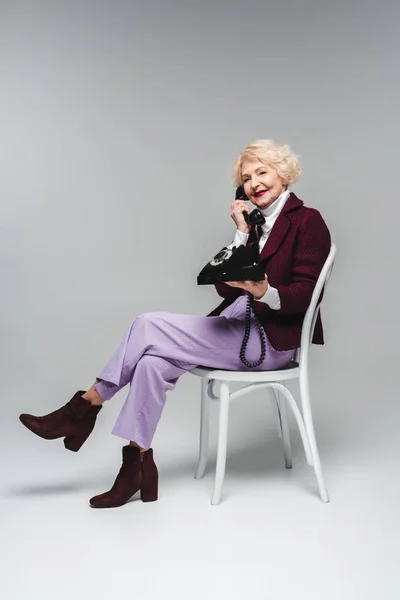 Beautiful senior woman talking by vintage phone while sitting on chair on grey — Stock Photo