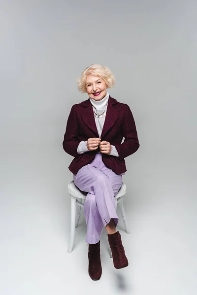 Красива старша жінка в стильній куртці сидить на стільці і дивиться на камеру на сірому — стокове фото