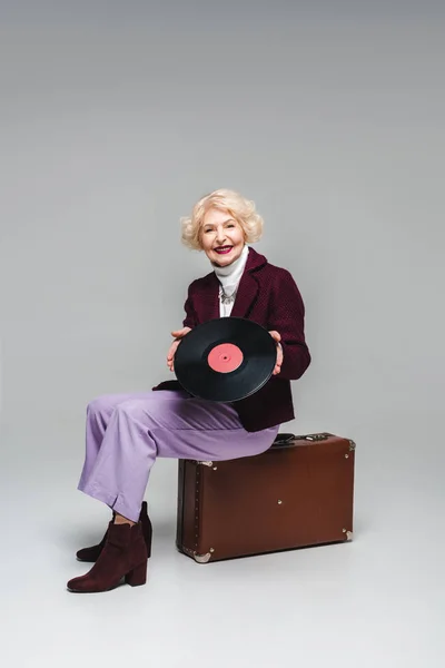 Happy stylish senior woman with vinyl disc sitting on vintage suitcase on gray background — Stock Photo