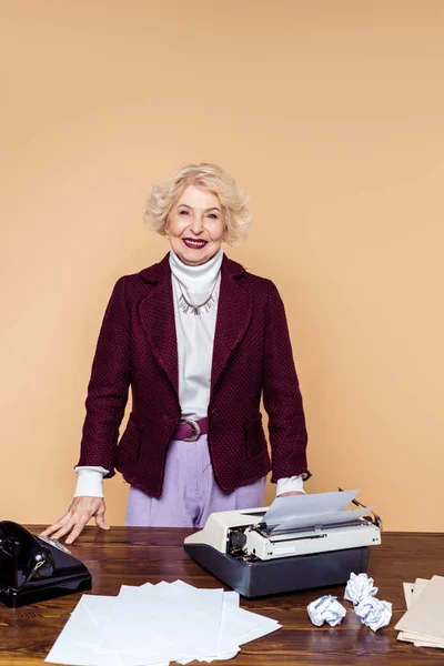 Улыбающаяся стильная пожилая женщина, стоящая за столом с печатной машинкой и ротационным телефоном — стоковое фото