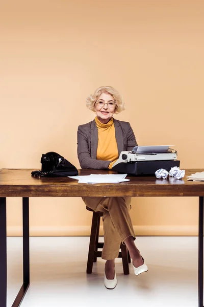 Усміхнена стильна старша жінка використовує друкарську машинку за столом з паперами та роторним телефоном — стокове фото