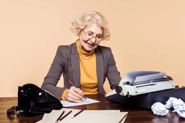 Stylish senior woman writing on paper at table with typewriter and rotary phone — Stock Photo