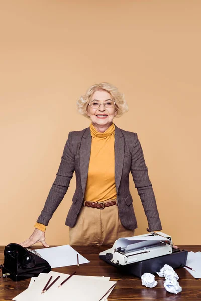 Sorrindo mulher idosa em óculos de pé à mesa com papéis, máquina de escrever e telefone rotativo — Fotografia de Stock