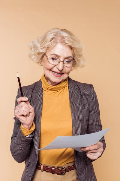 Felice donna anziana alla moda in occhiali con matita guardando la carta isolata su sfondo beige — Foto stock