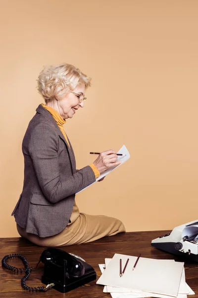 Seitenansicht einer stilvollen Seniorin, die auf Papier schreibt und mit Schreibmaschine und Drehtelefon auf dem Tisch sitzt — Stockfoto