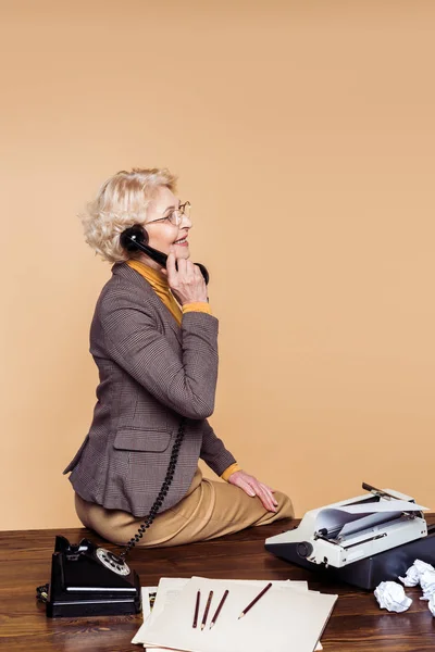Vue latérale de la femme âgée souriante parlant sur téléphone rotatif à table avec machine à écrire — Photo de stock