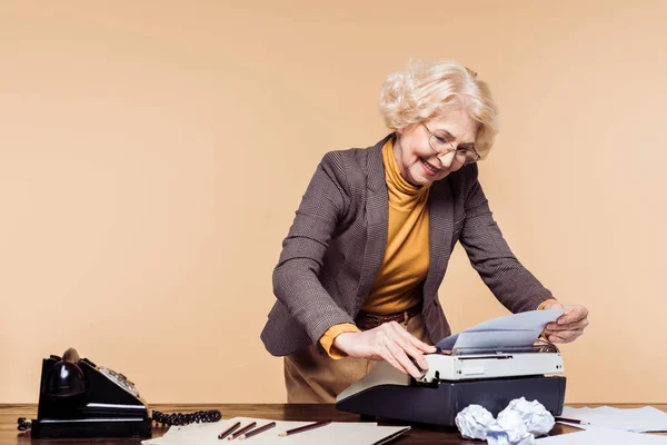 Mujer mayor sonriente usando máquina de escribir en la mesa con teléfono giratorio - foto de stock