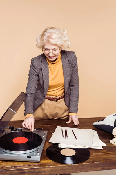 Senior stylish woman turning on vinyl record player at table with typewriter and vinyl disc — Stock Photo