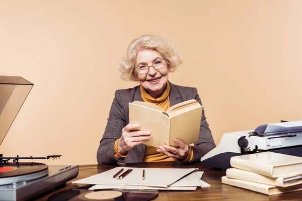 Усміхнена старша жінка в окулярах з книгою, що сидить за столом з вініловим диском, програвачем і друкарською машинкою — стокове фото