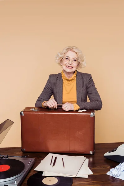 Счастливая стильная пожилая женщина с винтажным чемоданом за столом с виниловым диском, проигрывателем и пишущей машинкой — стоковое фото