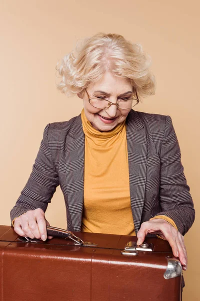Fashionable senior woman in eyeglasses closing vintage suitcase isolated on beige background — Stock Photo
