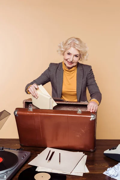Стильная пожилая женщина кладет книги в винтажный чемодан на стол с магнитофоном и виниловым диском — стоковое фото