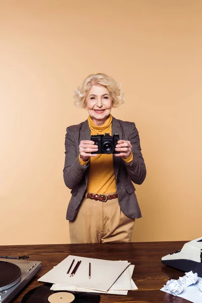 Lächelnde stilvolle Seniorin mit Filmkamera auf beigem Hintergrund — Stockfoto