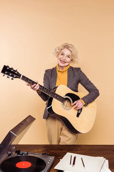 Mulher sênior sorrindo tocando na guitarra acústica perto da mesa com leitor de vinil — Fotografia de Stock