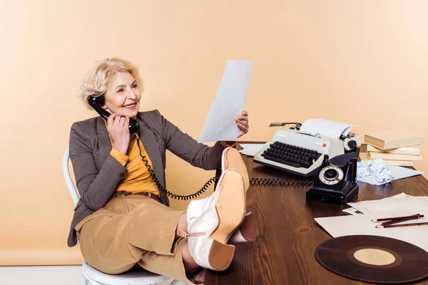 Donna anziana sorridente con le gambe sul tavolo che parla al telefono rotante e guarda pape — Stock Photo