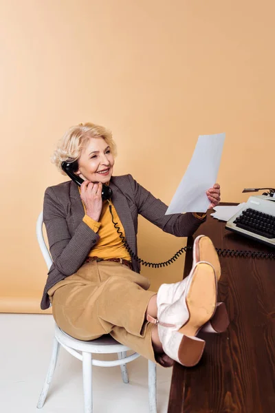 Élégante femme âgée avec les jambes sur la table parlant sur téléphone rotatif et regardant le papier — Photo de stock