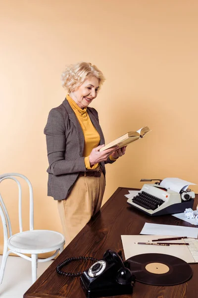Sonriente mujer mayor lectura libro cerca de la mesa con teléfono giratorio, disco de vinilo y máquina de escribir - foto de stock